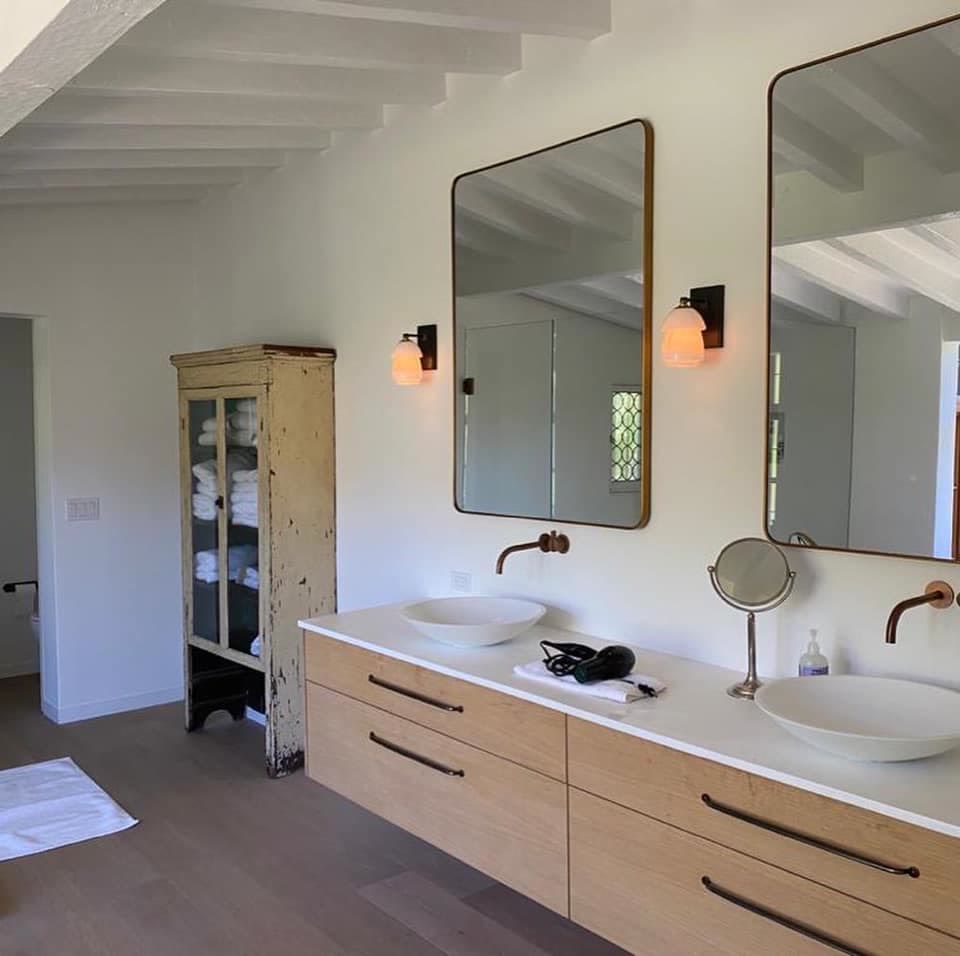 A bathroom with a white beamed ceiling, white walls, a double oak vanity with two vessel basins, brass-framed mirrors and an old distressed cabinet holding towels.