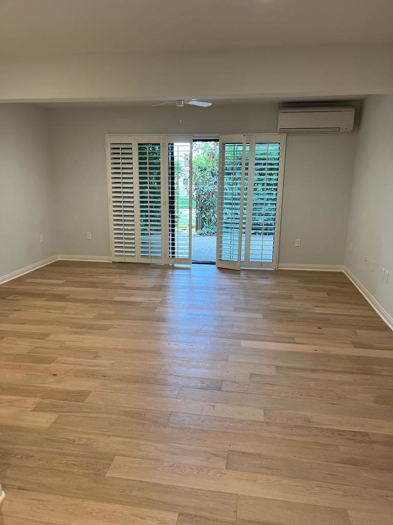 An empty room with gray walls, white baseboards, a light wood floor and white plantation shutters opening onto a garden.