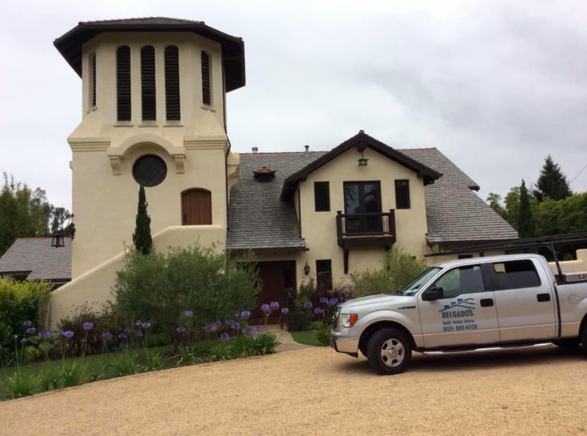 A silver pickup lettered Delgado's Quality Painting Services with the phone number (805) 689-4528, parked on a gravel drive in front of a cream stucco house with a bell tower.